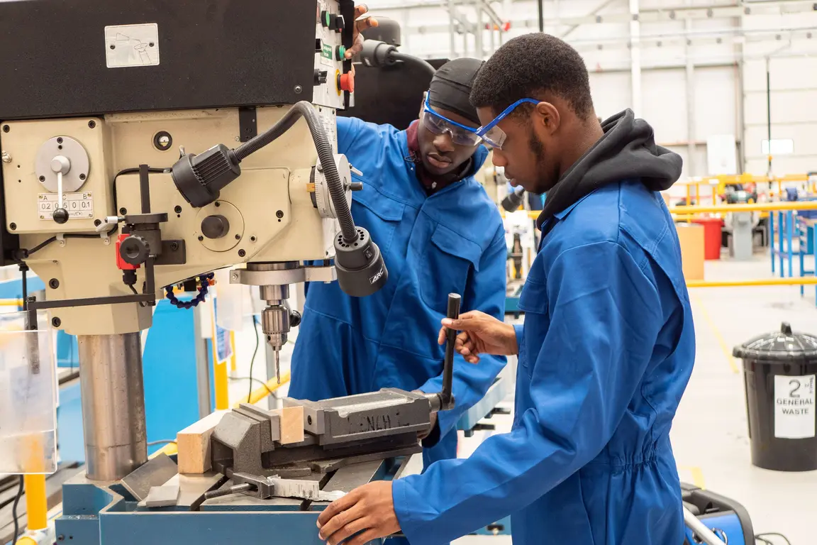 Two men wearing blue jumpsuits and goggles, one holding a tool, working on a drill press in a factory.