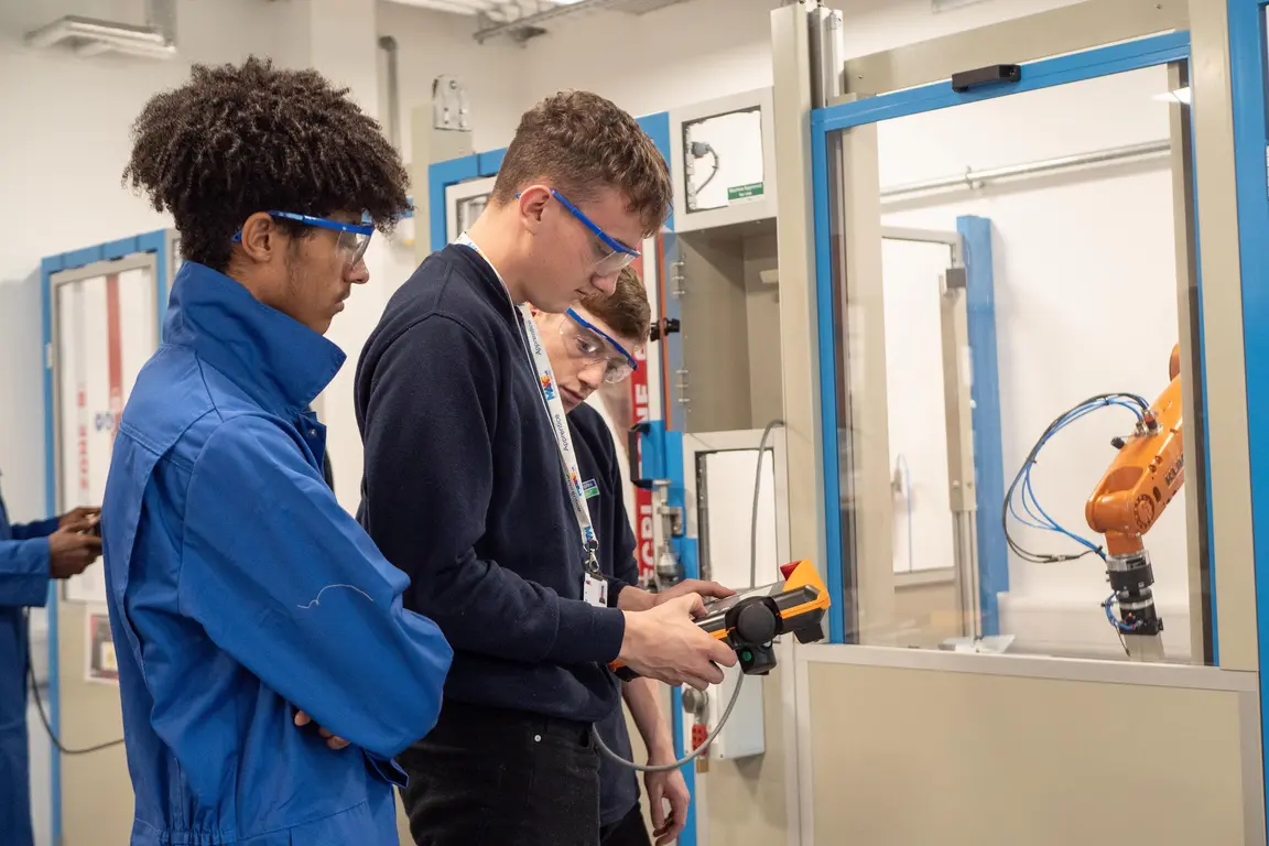 Three students in blue jumpsuits examine a robotic arm with a laser scanner.