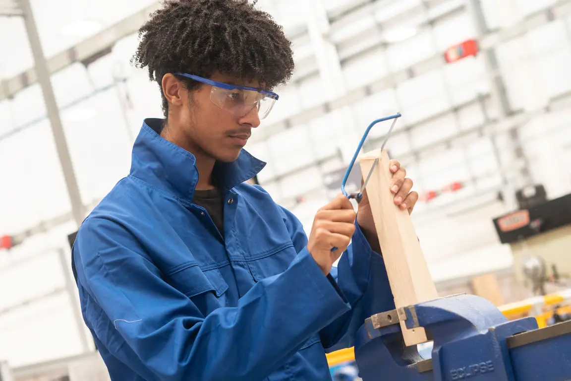 Apprentice in blue jacket working in a shop, wearing goggles and holding a wooden plank.