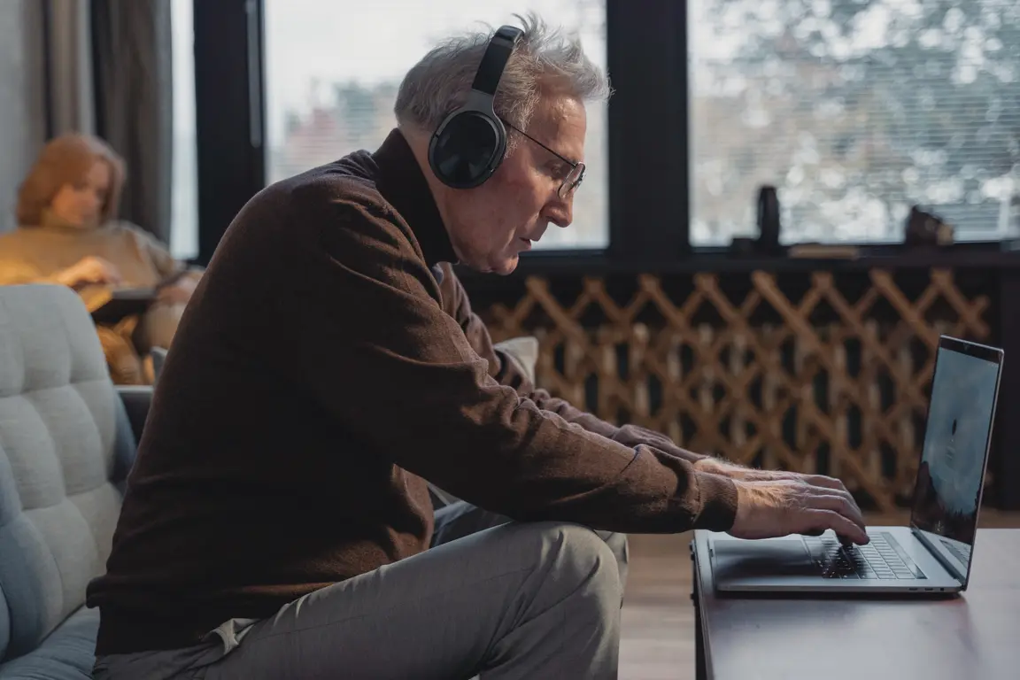 an older man with headphones on sitting forward engaged with his laptop