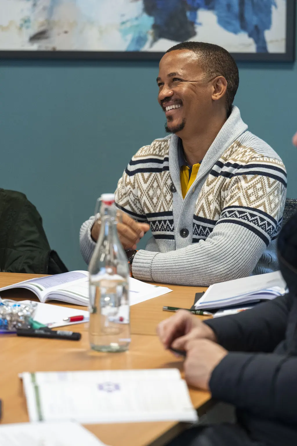 Three men seated around a conference table, two with papers, one smiling, one with glasses.