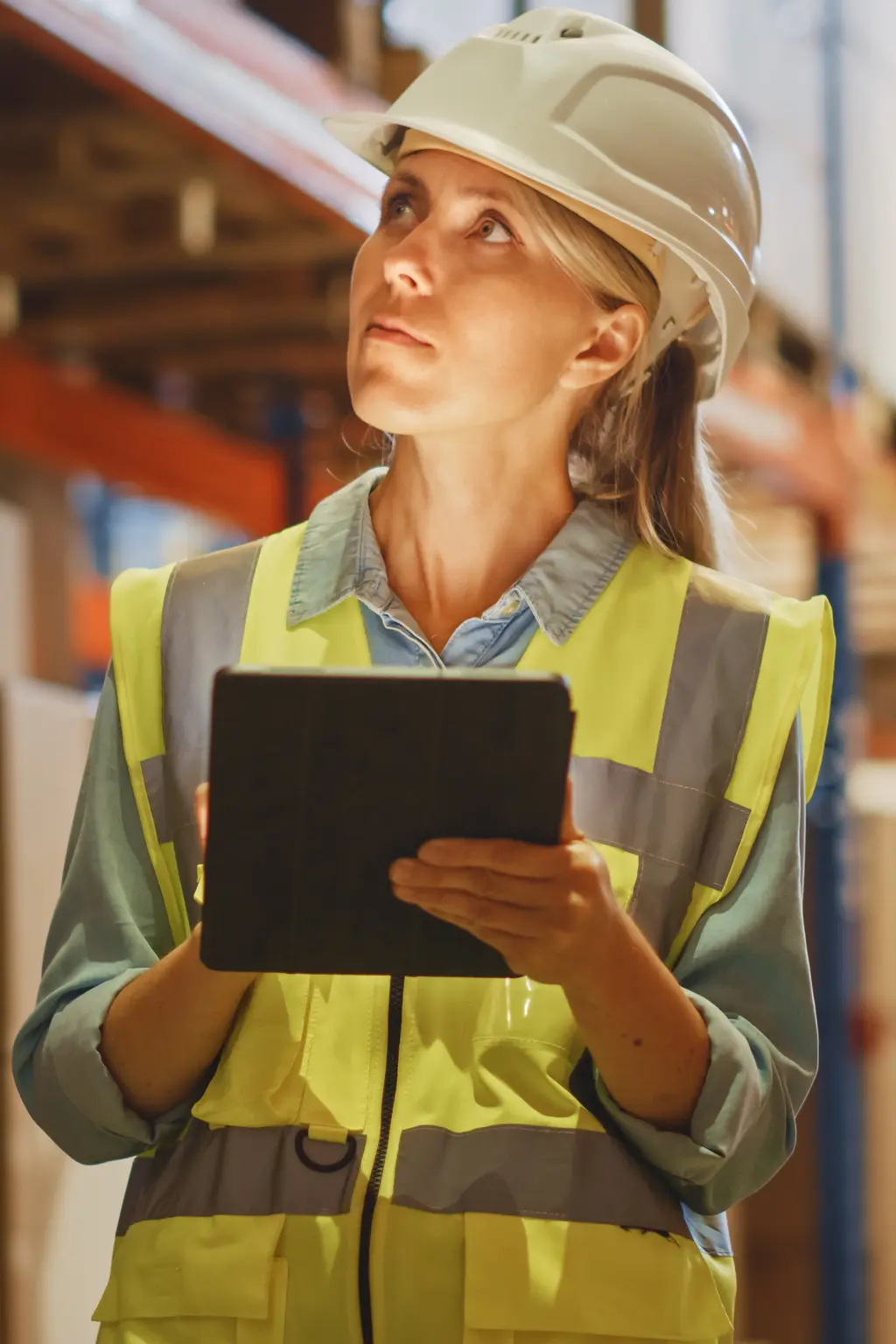 A woman in a yellow safety vest and hard hat holds a tablet in a warehouse.