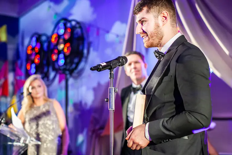 Man with make uk award in black suit standing on stage in front of microphone.