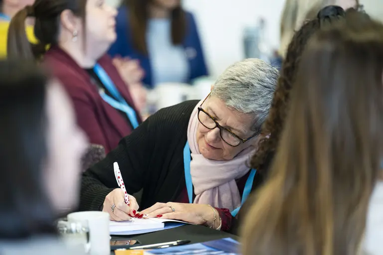 A woman wearing glasses and scarf is writing on a notepad at a conference.