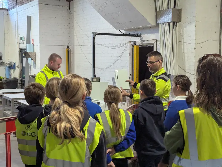 Children wearing yellow vests listen to a man explain something in a factory on national manufacturing day.