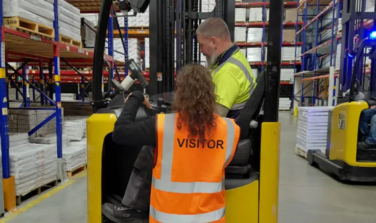 A woman in a yellow vest reading "VISITOR" instructs a man on a forklift in a warehouse.