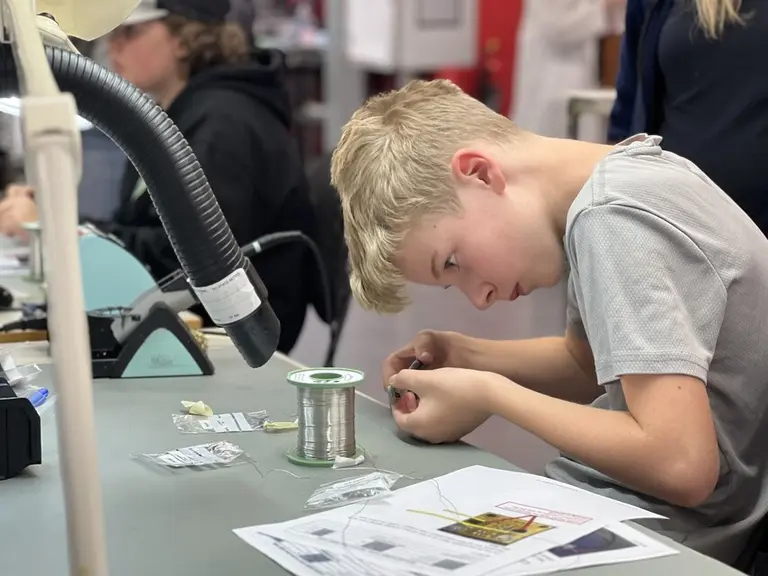 A boy in a gray shirt intently working with small wires and components at a workbench.