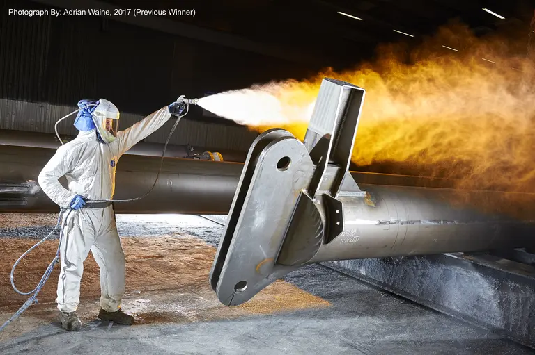 Worker in white jumpsuit sprays torch flame on metal part.