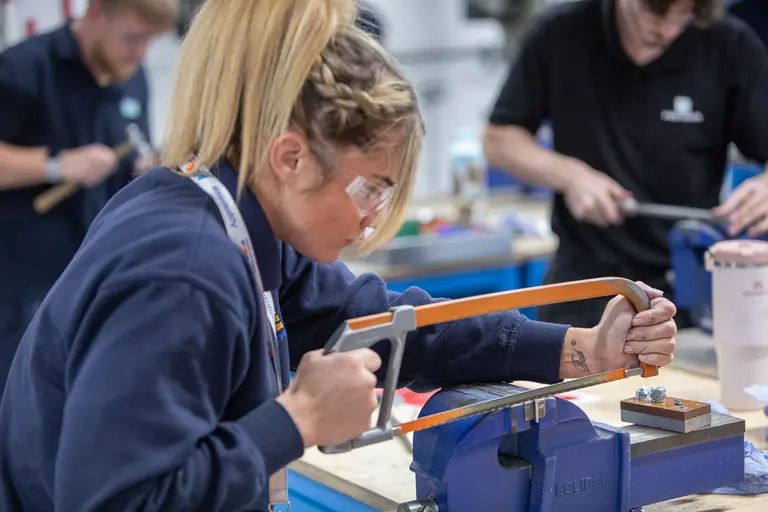 Woman wearing safety glasses saws metal with a hacksaw.